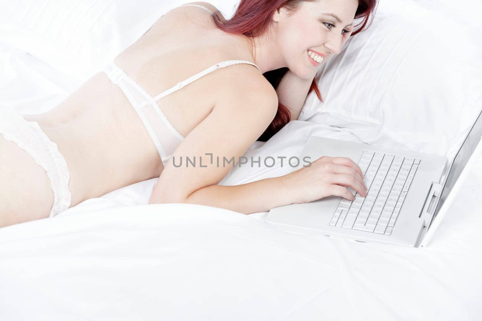Attractive young woman working on her laptop in white underwear on her bed at home