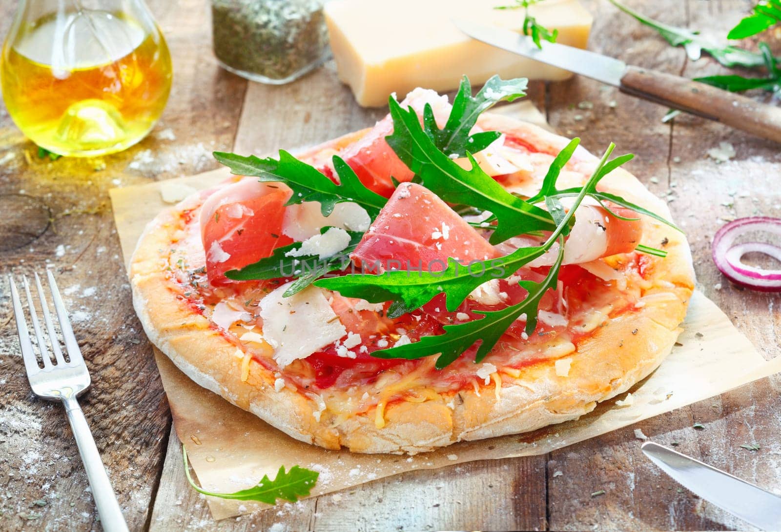 Preparing a tasty pizza in the kitchen using a crisp base topped with fresh herbs, ham, rucola, cheese and tomato