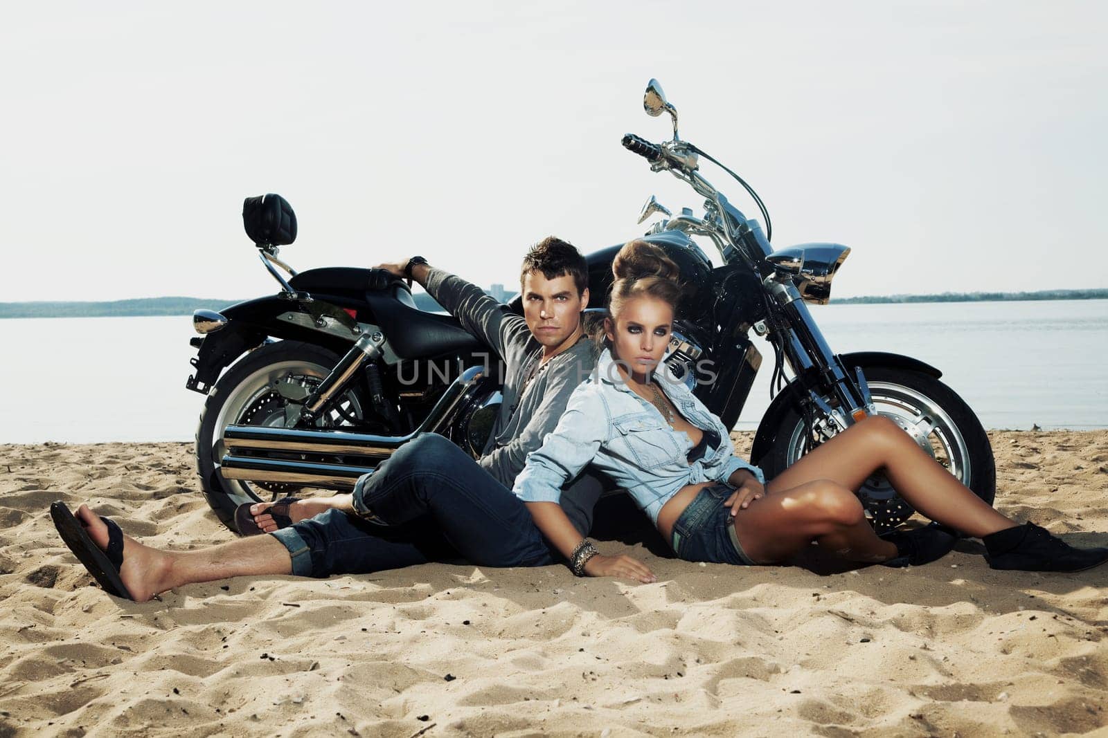 Young handsome boyfriend and girlfriend riders sitting together on sand beach by beautiful bike - travel
