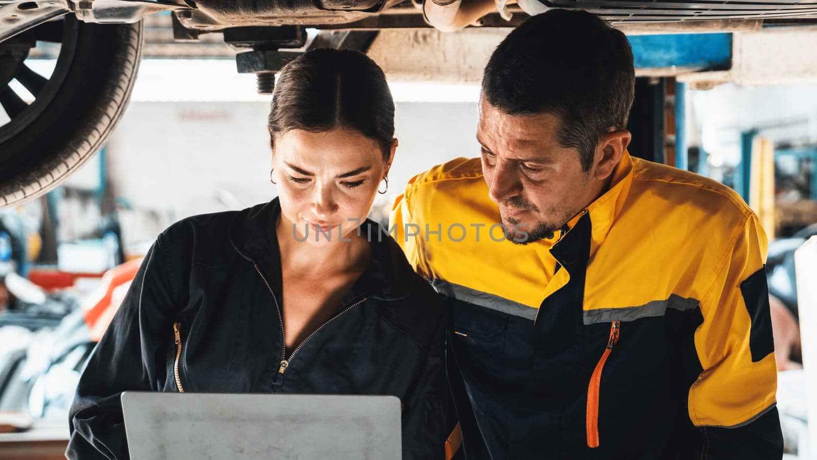 Two vehicle mechanic working together underneath lifted car, conduct car inspection with laptop. Automotive service technician in uniform carefully make diagnostic troubleshooting. Panorama Oxus