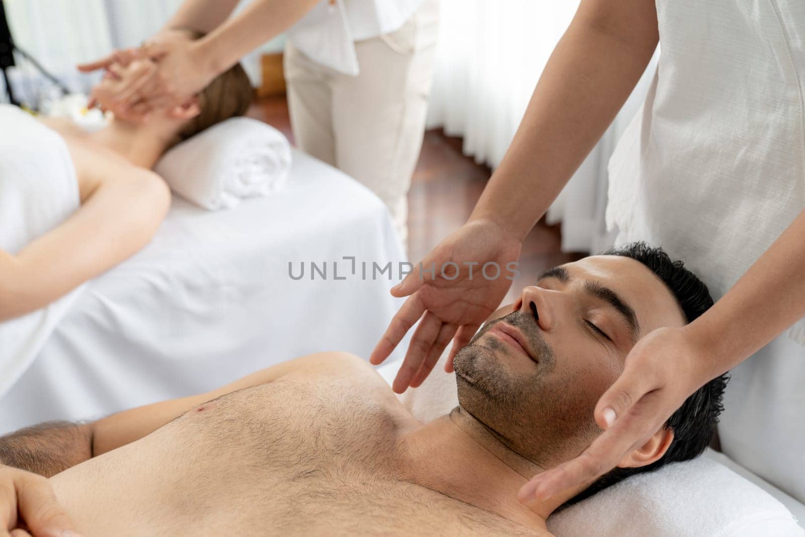 Caucasian couple enjoying relaxing anti-stress head massage and pampering facial beauty skin recreation leisure in dayspa modern light ambient at luxury resort or hotel spa salon. Quiescent