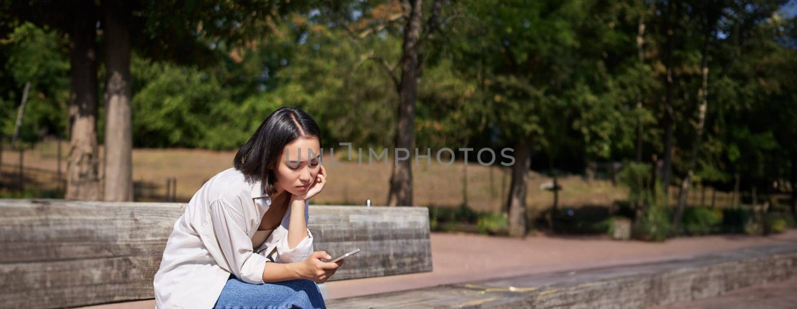 Portrait of asian girl sitting with smartphone feeling sad, looking gloomy and frustrated, waiting for a call outdoors in park.