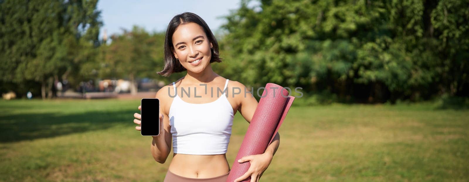 Smiling asian fitness girl with rubber yoga mat, shows her smartphone screen, recommends workout application, stands on lawn in park by Benzoix