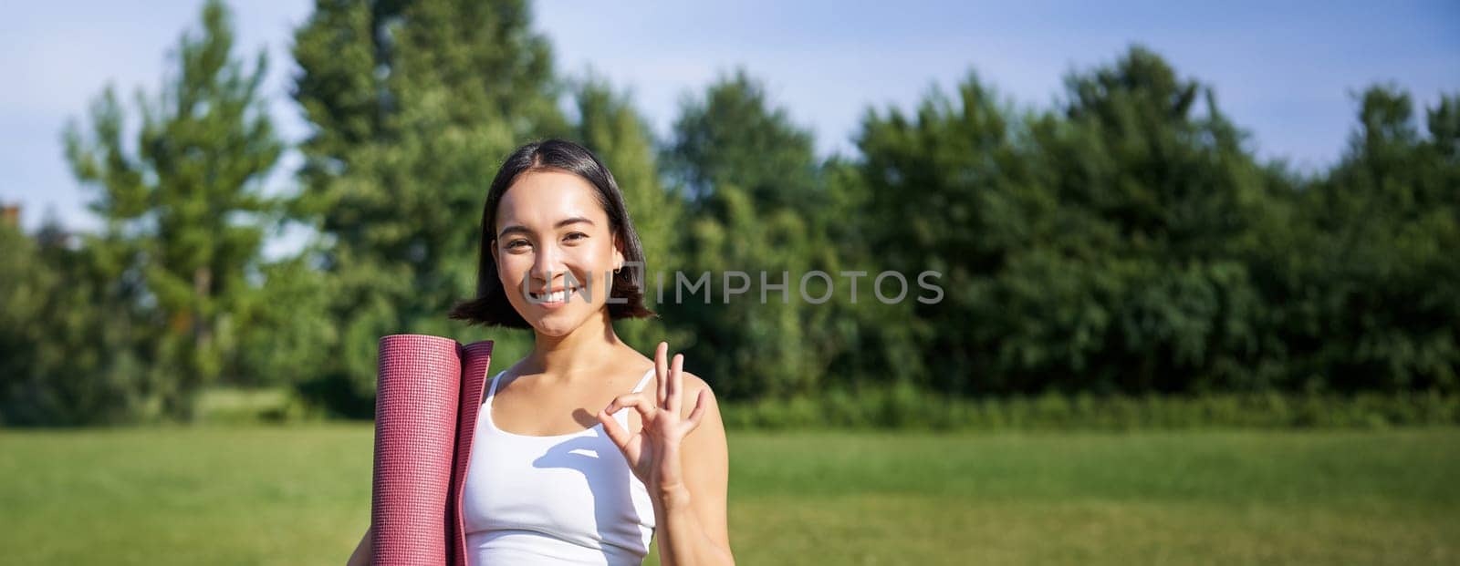 Excited young woman standing with sports mat, yoga clothes, shows okay sign, workout in park, wellbeing training session outdoors by Benzoix