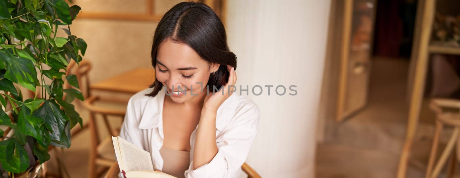 Vertical shot of happy young asian woman enjoys reading, sitting with book in cafe, drinking coffee and eating croissant by Benzoix