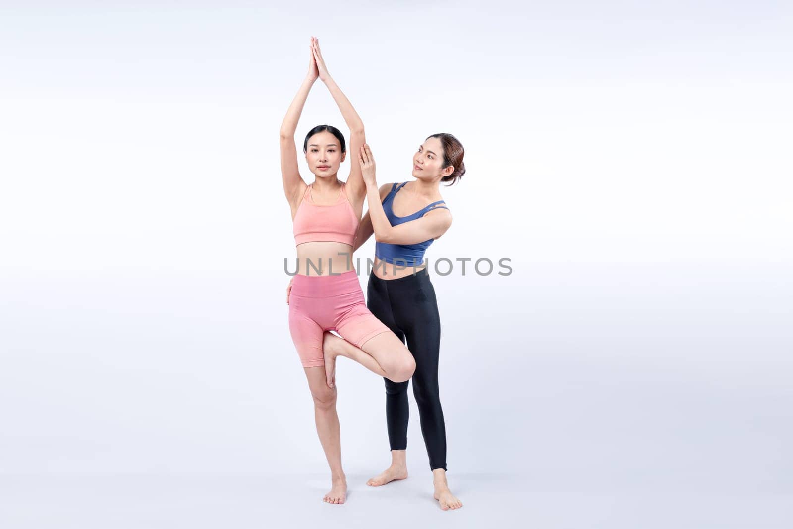 Asian woman in sportswear doing yoga exercise in standing pose with her trainer or yoga buddy. Healthy body care and meditation yoga lifestyle in full shot on isolated background. Vigorous