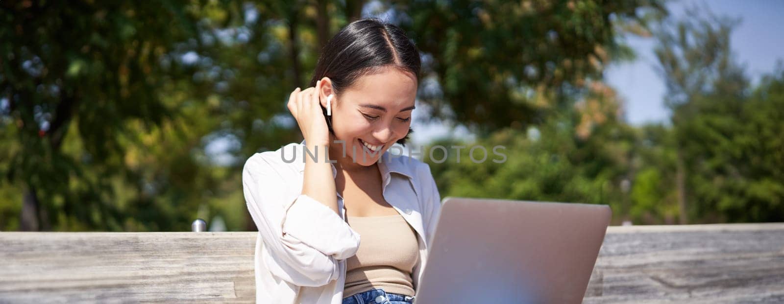 Beautiful asian girl laughing, watching video on laptop, listening music or video chatting online while sitting in park on bench.