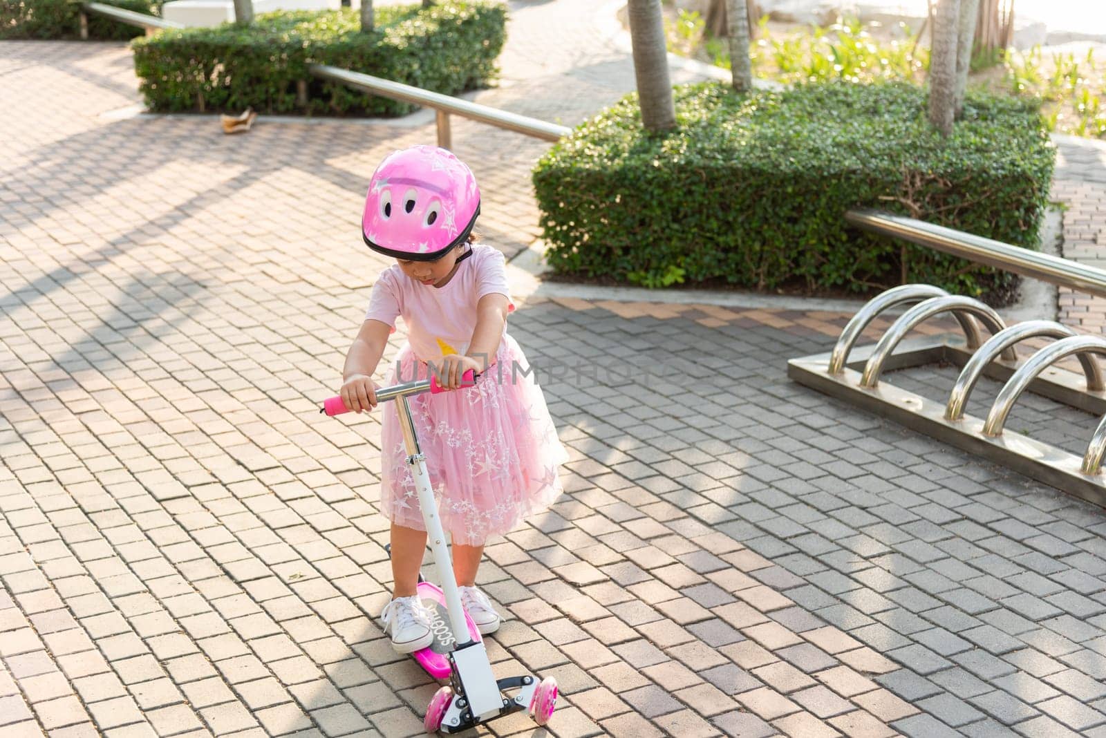 Happy Asian little kid girl wear safe helmet playing pink kick board on road in park outdoors on summer day by Sorapop