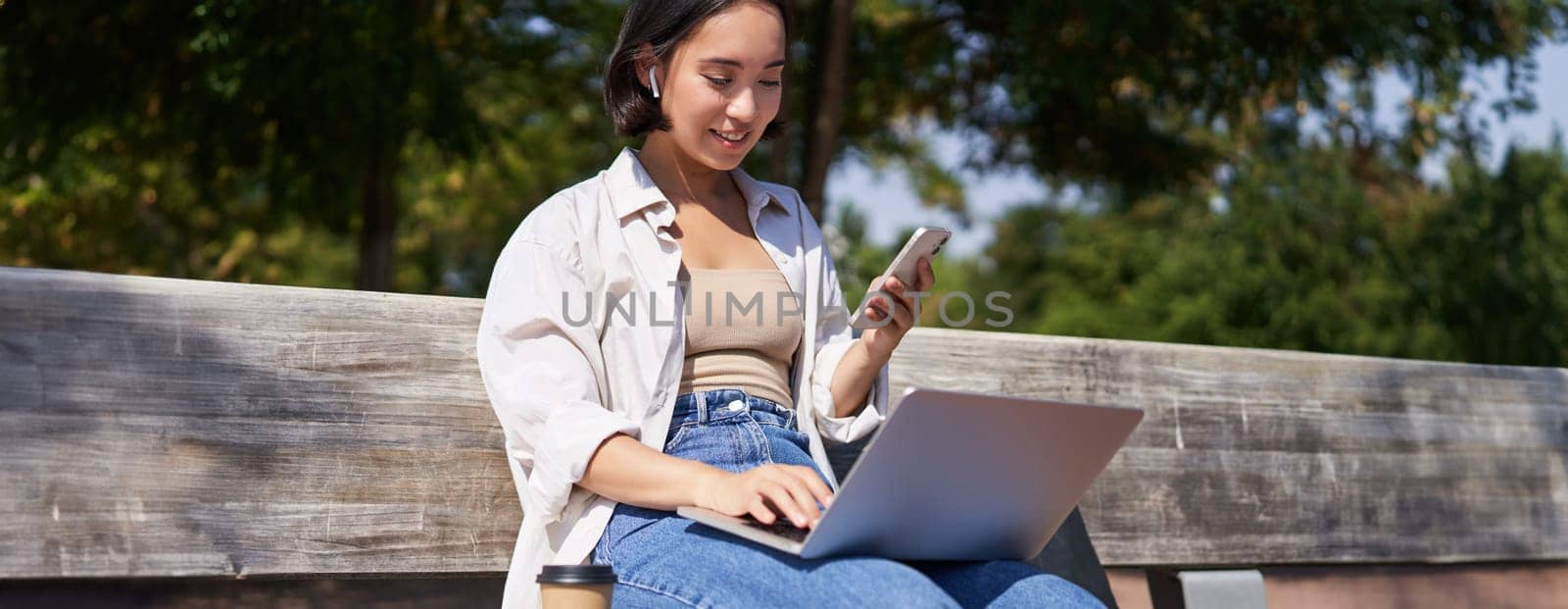 Young asian girl talking on mobile phone, using laptop, working online from park, sitting on bench on sunny day with computer.