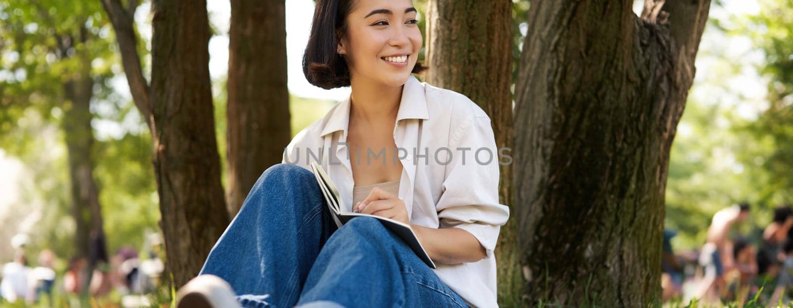 Portrait of happy asian girl sitting in park and writing in her diary. Young woman doing homework on fresh air, sitting near tree and smiling.