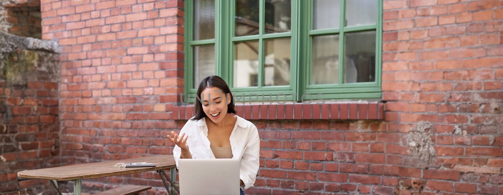 Vertical shot of asian girl with laptop, talking on video call, chatting online, sitting in cafe outdoors. People and communication concept