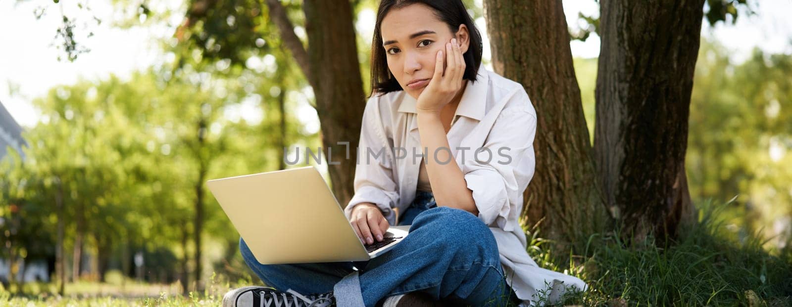 Portrait of young asian woman sitting in park near tree, working on laptop, using computer outdoors by Benzoix