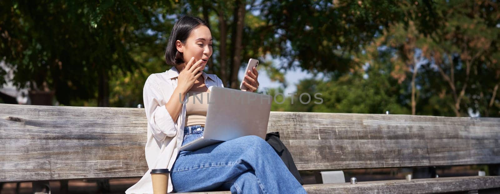 Asian girl looks surprised at smartphone screen, checking mobile phone notifications and looking excited, sitting with laptop in park on bench.