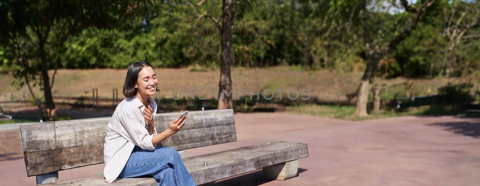 Stylish young asian girl, student sitting in park with smartphone, using telephone, waiting for someone while sitting on bench by Benzoix