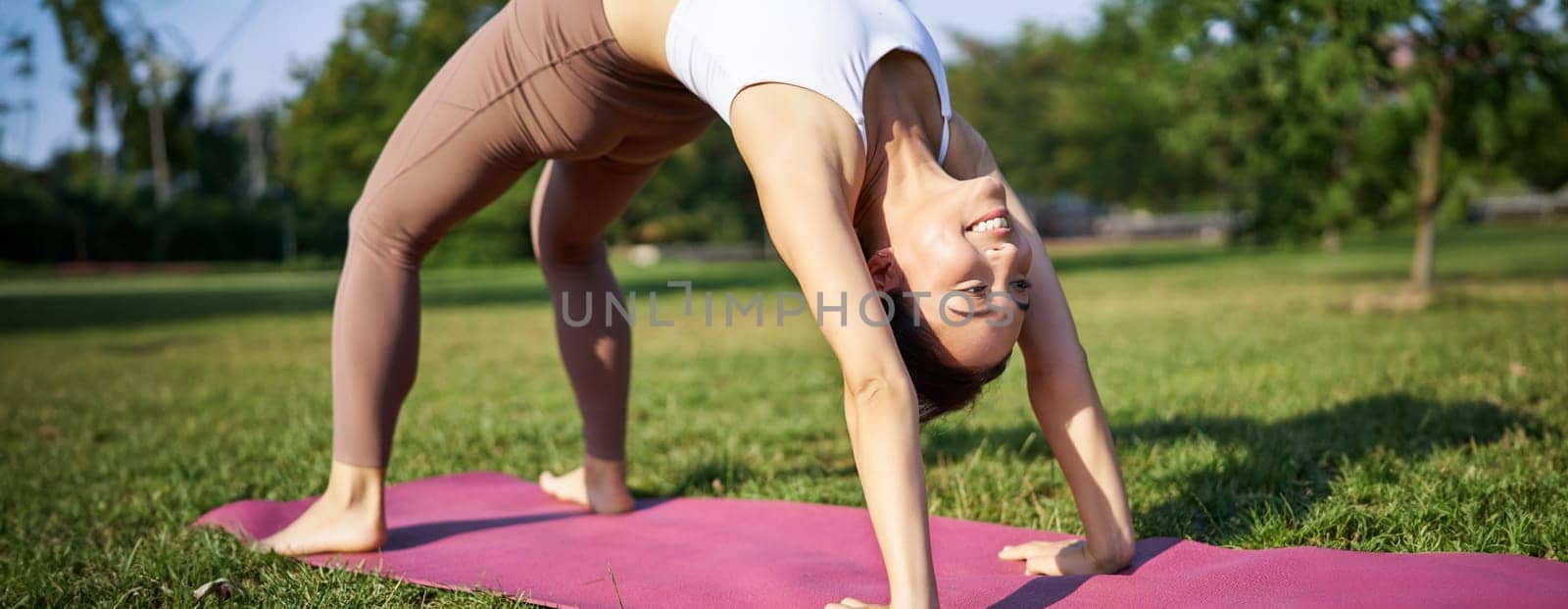 Woman smiling while making bridge asana, doing yoga in park on rubber mat by Benzoix