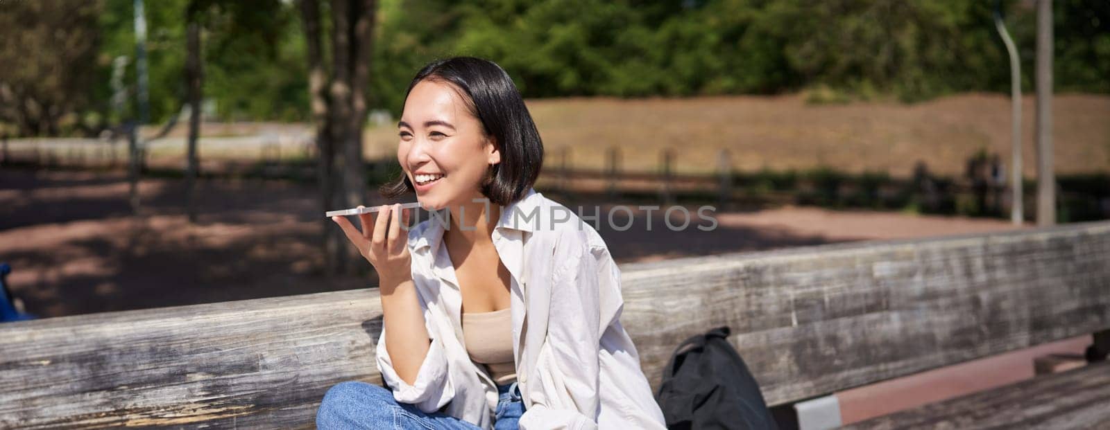 Portrait of happy asian girl records voice message, talking via speakerphone on smartphone, sitting relaxed on bench in park on sunny day by Benzoix