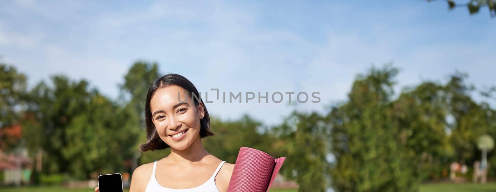 Excited fitness girl recommends application for sport and workout, shows phone screen, standing with rubber yoga mat in park after training session.