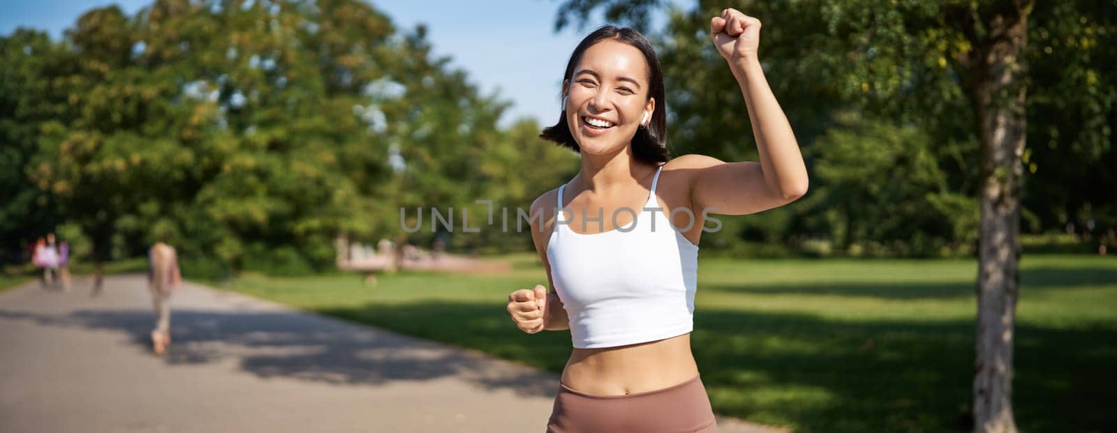 Happy fitness girl achieve goal, finish marathon, running with hands up, celebrating victory while jogging, triumphing in park.
