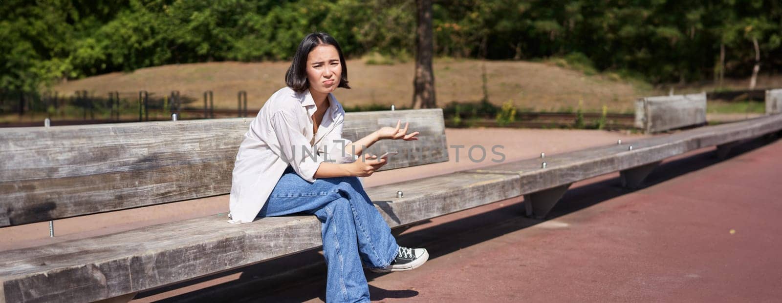 Depressed asian girl sits on bench in park with smartphone, feeling uneasy and stressed, frowning and sighing by Benzoix