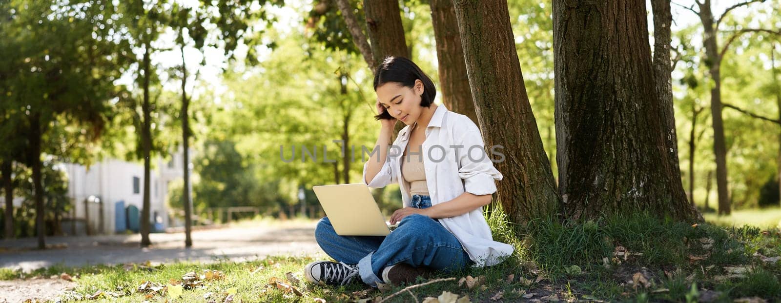 Remote work. Smiling asian girl, student doing homework remotely from park, sitting with laptop near tree and typing.