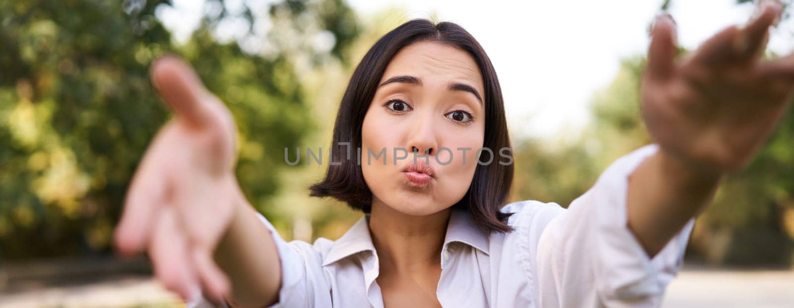 People and mobile connection. Happy young asian woman takes selfie on smartphone, holds camera with hands, poses in park on summer day by Benzoix