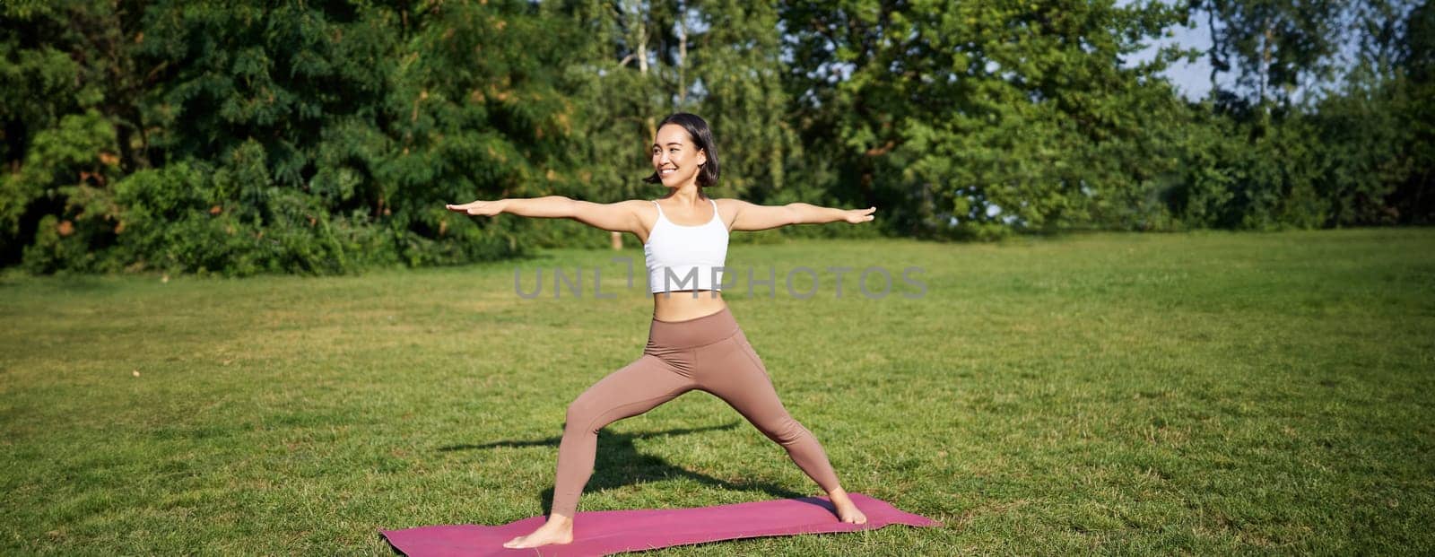Young woman stretching her body, workout and doing yoga in park, standing on rubber mat in fitness clothing during training session.