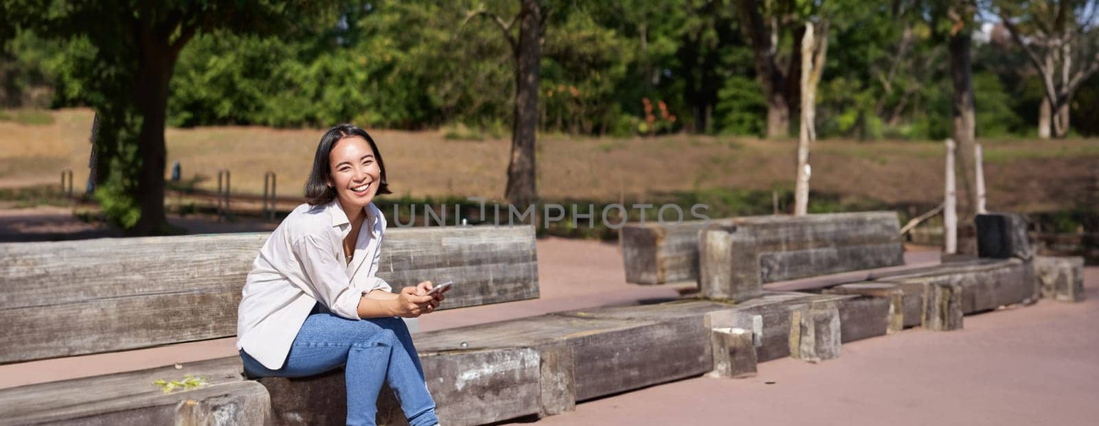 Happy smiling asian girl sitting on bench in park and browsing on social media, holding smartphone, using mobile phone app.
