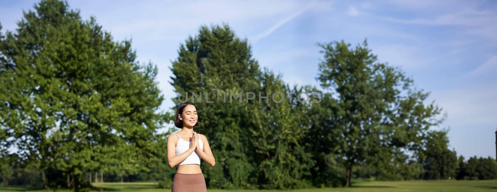 Young mindful woman, meditating on fresh air, doing yoga exercises in park, standing on rubber mat in sportswear.