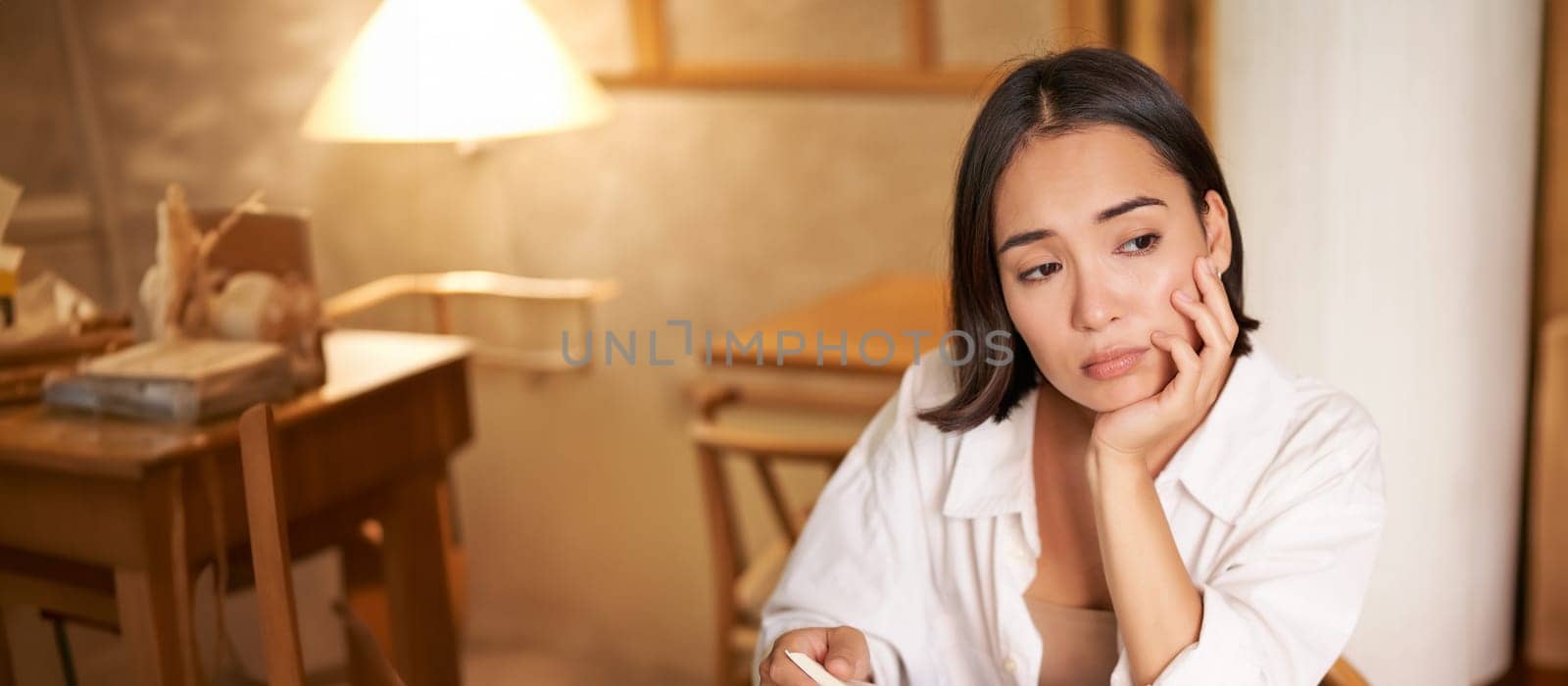 Vertical shot of asian girl sits alone in cafe, reads book and looks upset, drinks coffee with croissant.