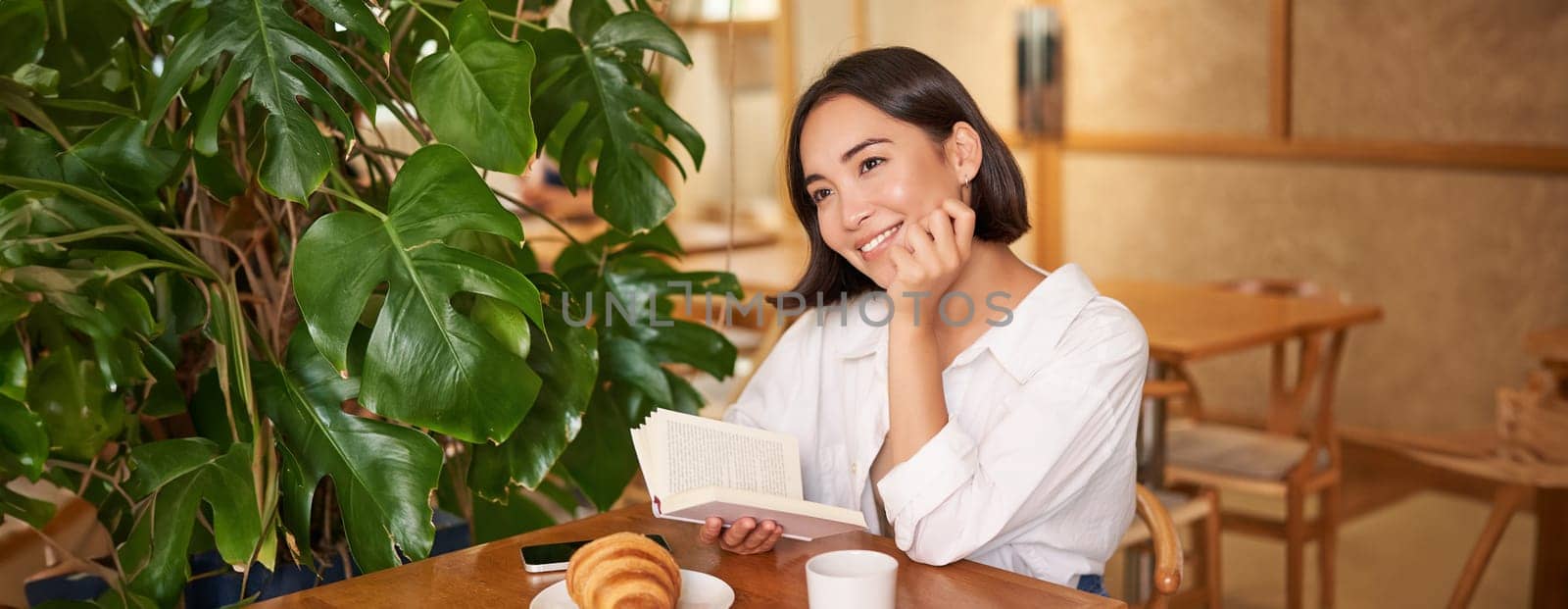 Dreamy young smiling asian woman reading book, sitting in cafe, eating croissant and drinking coffee in cozy interior.