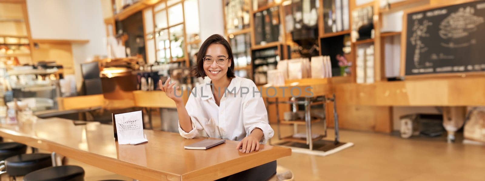 Young stylish asian woman, business owner in glasses, sitting in cafe with notebook, smiling at camera. People and lifestyle concept