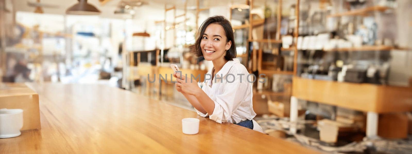 Vertical portrait of stylish asian woman sitting in cafe, drinking coffee and using smartphone. Lifestyle and people concept
