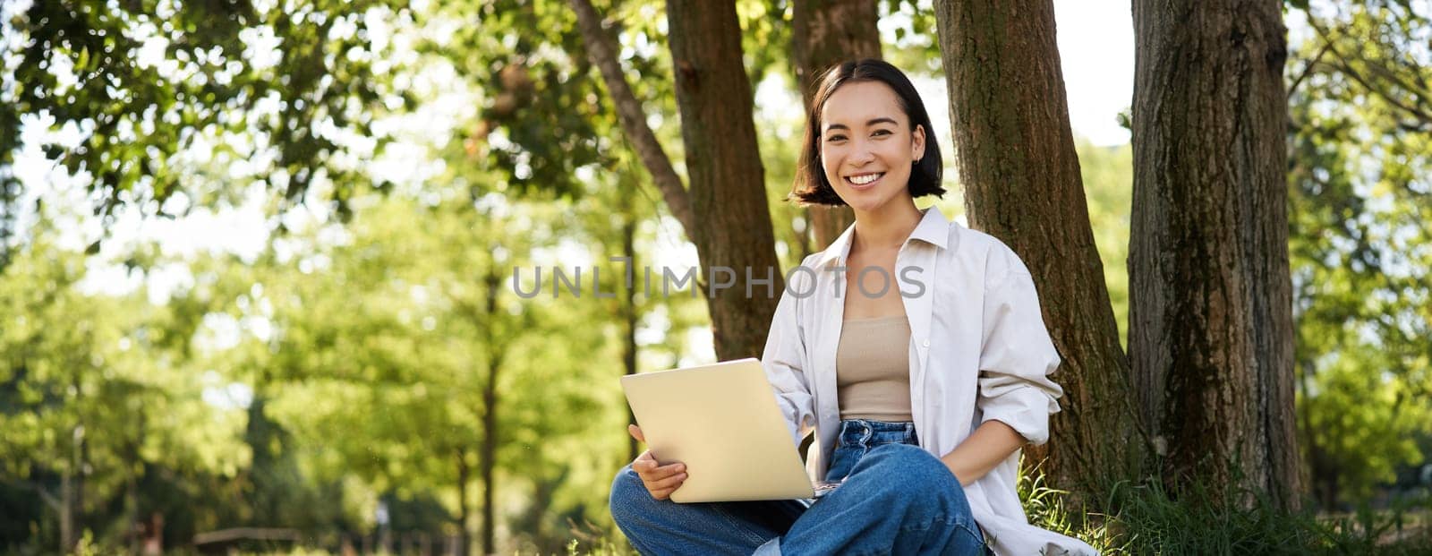 Portrait of asian girl with laptop, sitting near tree in park on sunny summer day, working on remote, e-learning with computer.