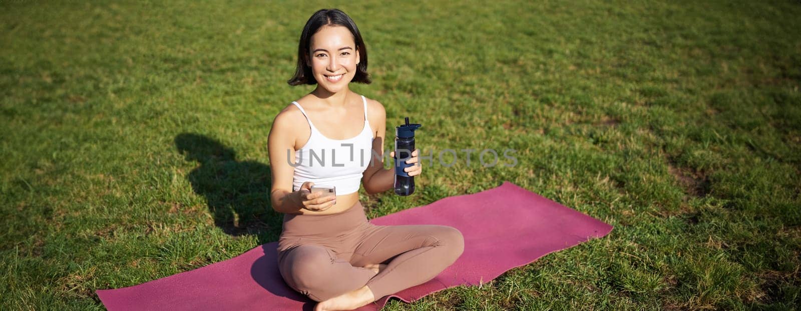 Smiling young asian woman sits on grass, doing exercises in park, workout on lawn, drinks water from bottle.
