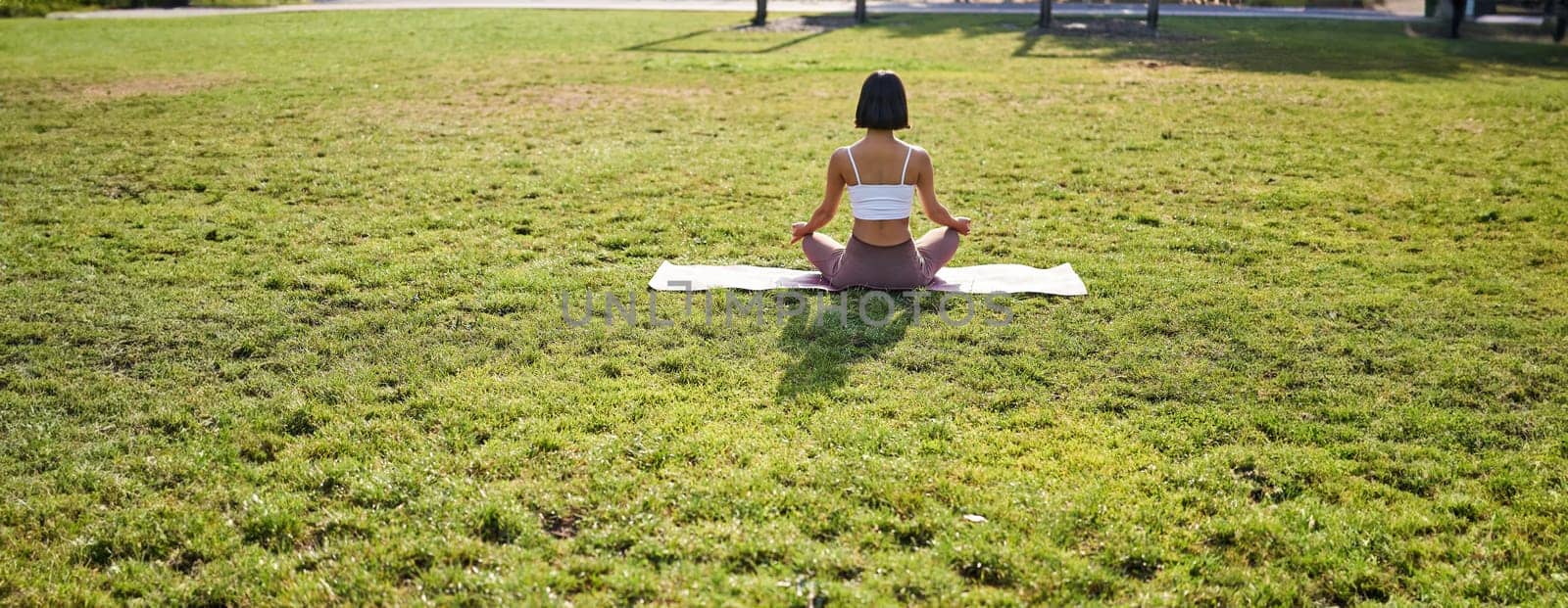 Rear view of young woman meditating in morning park, relaxing, breathing practice and wellbeing training session.