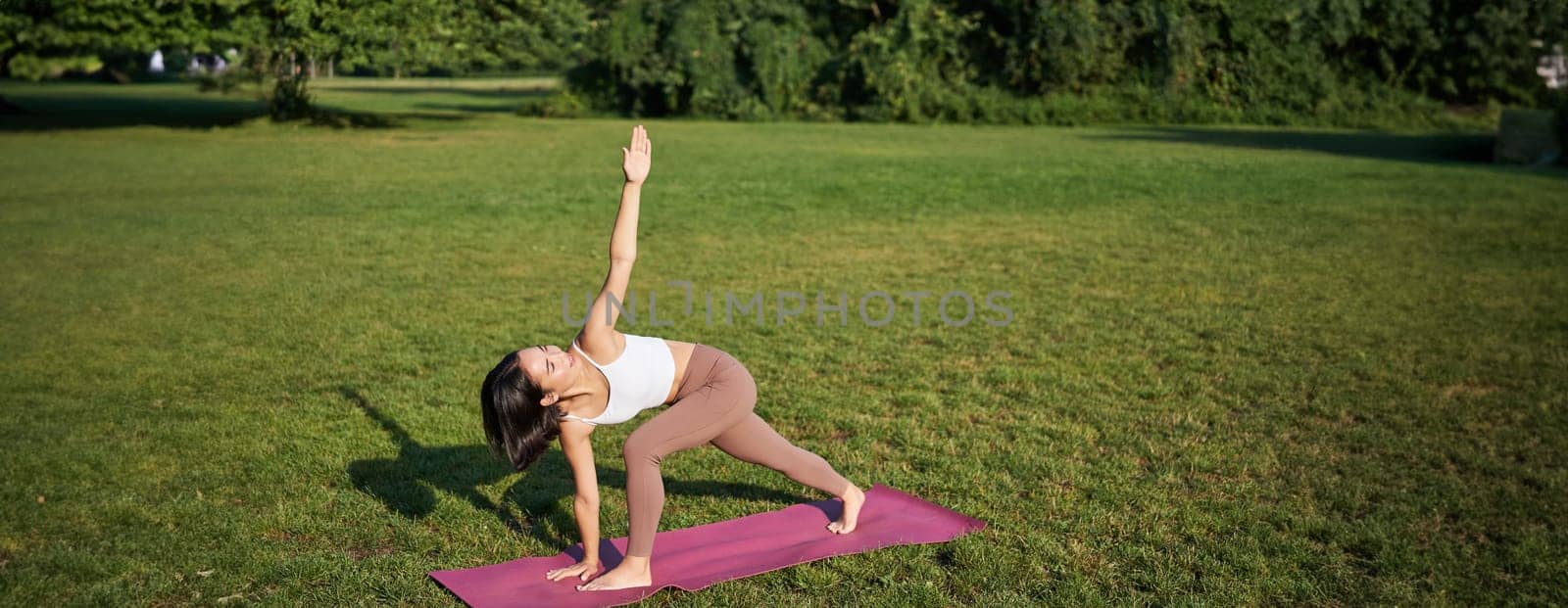 Portrait of young asian woman stretching, doing yoga on rubber mat, exercising in park, mindful training on fresh air.