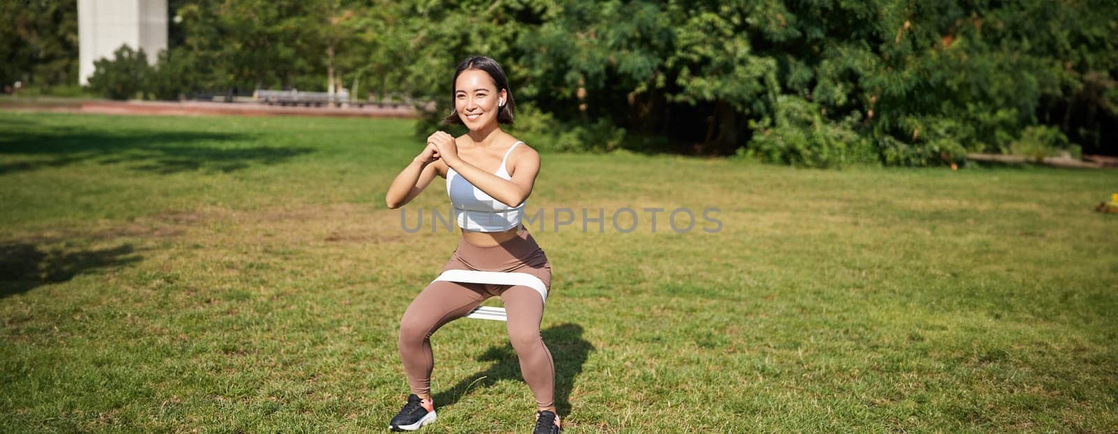 Woman using stretching band, resistance fitness rope for workout in park, doing squats, training on fresh air by Benzoix