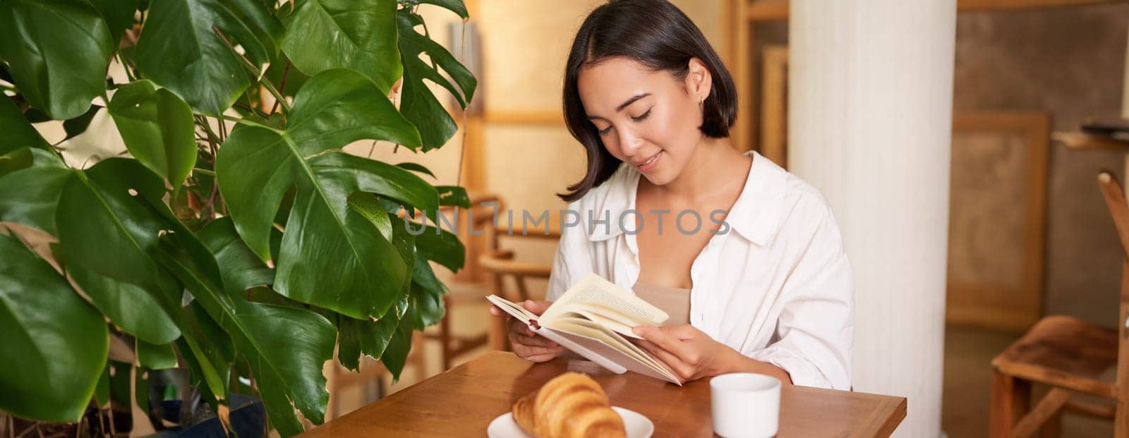 Smiling young asian woman enjoys reading book while sits alone in cafe, drinks coffee, looking at page by Benzoix