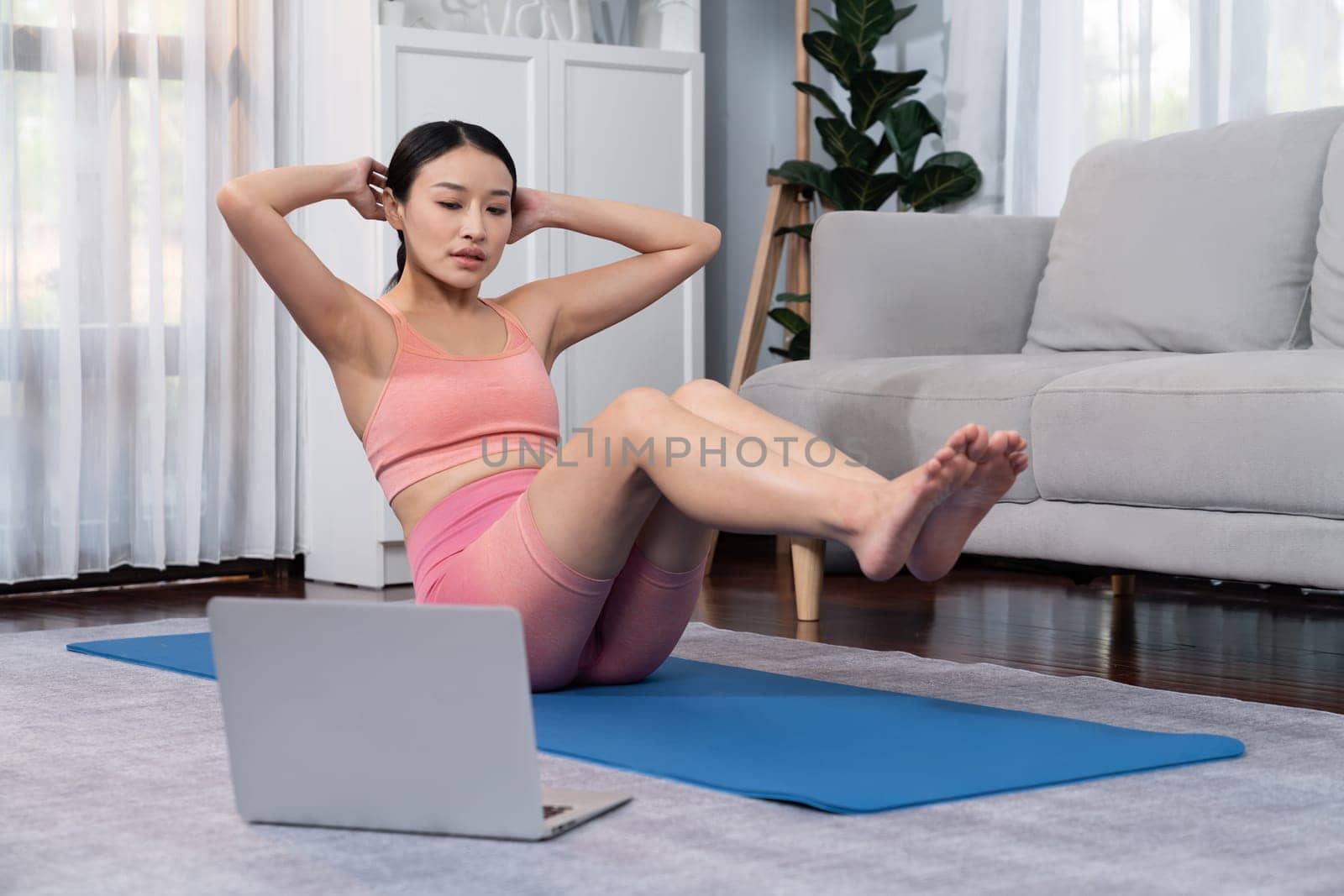 Asian woman in sportswear doing crunch on exercising mat as home workout training routine. Attractive girl engage in her pursuit of healthy lifestyle with online exercise training video. Vigorous