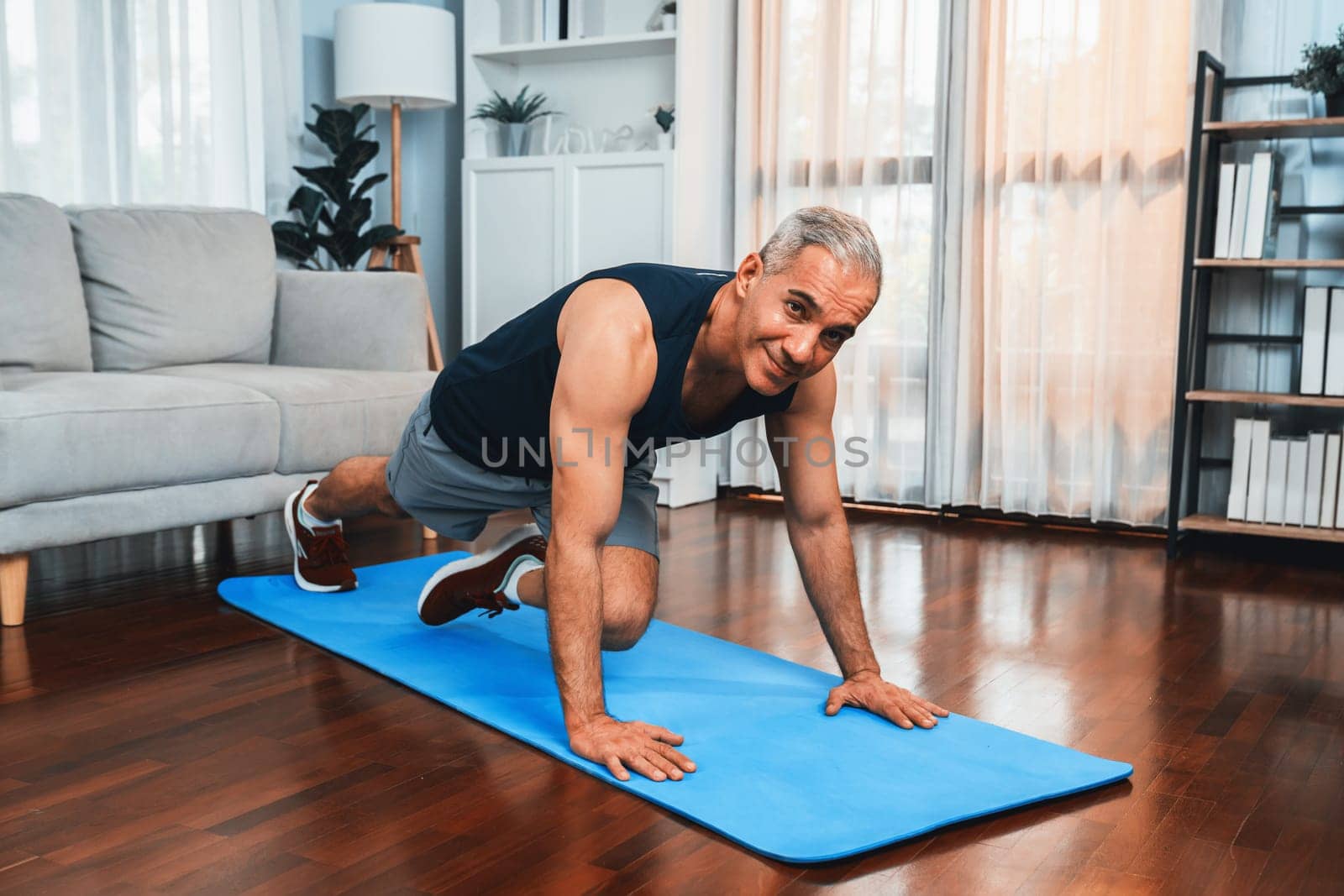 Athletic and active senior man doing exercise on fit mat with plank climbing at home exercise as concept of healthy fit body lifestyle after retirement. Clout