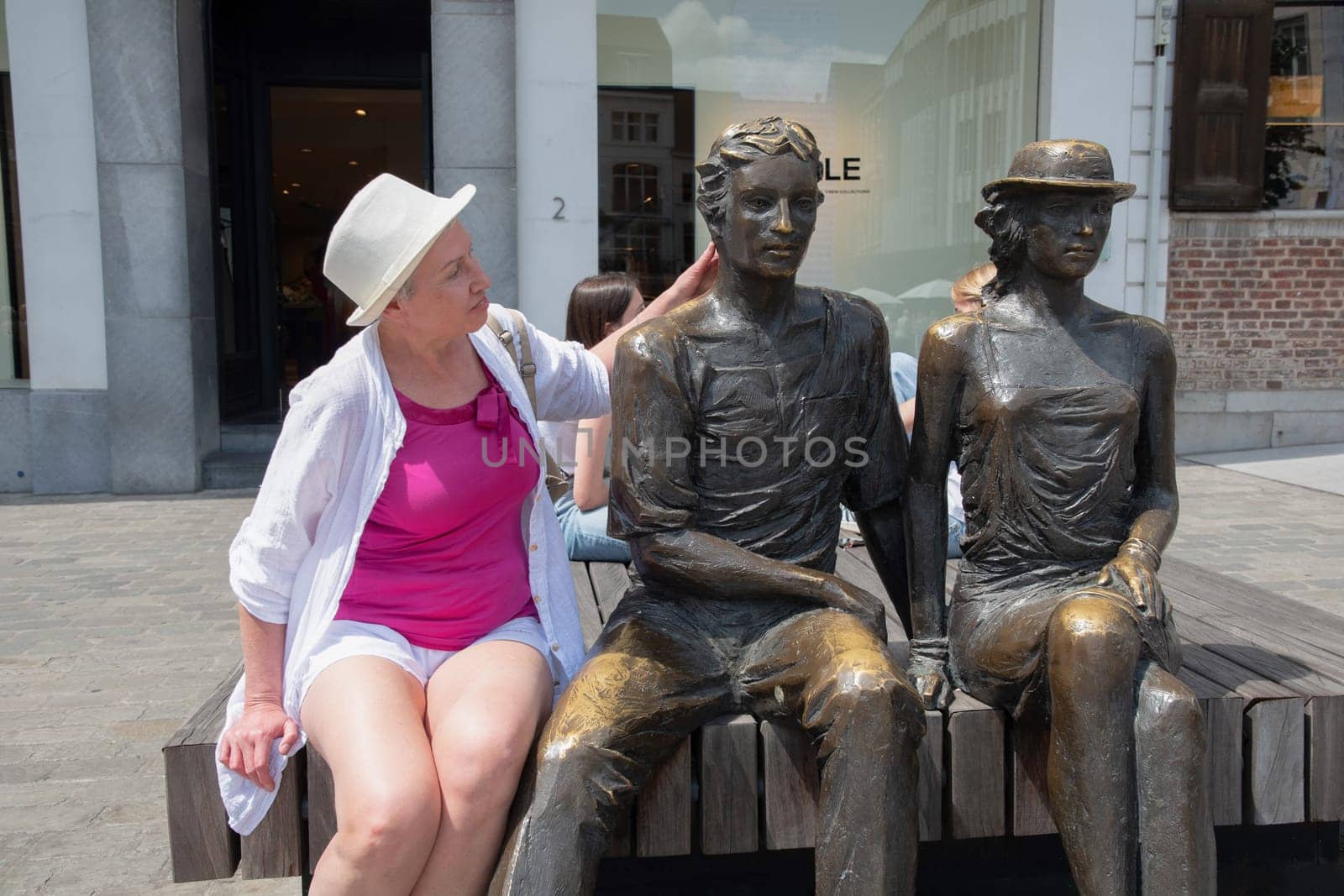 A middle-aged woman sits on a bench with two bronze statues Hendrik and Katrien, Sculpture by artist Marc Cox with the official title Listening to the carillon, Hasselt, Belgium, July 8, 2023