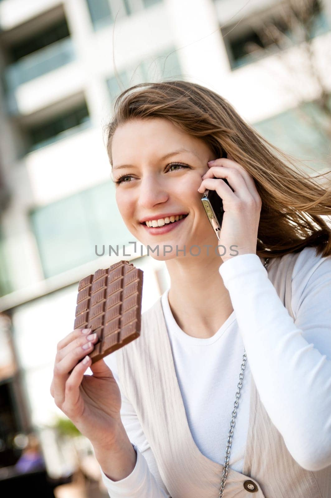 Outdoor portrait of young smiling woman eating chocolate calling with mobile phone