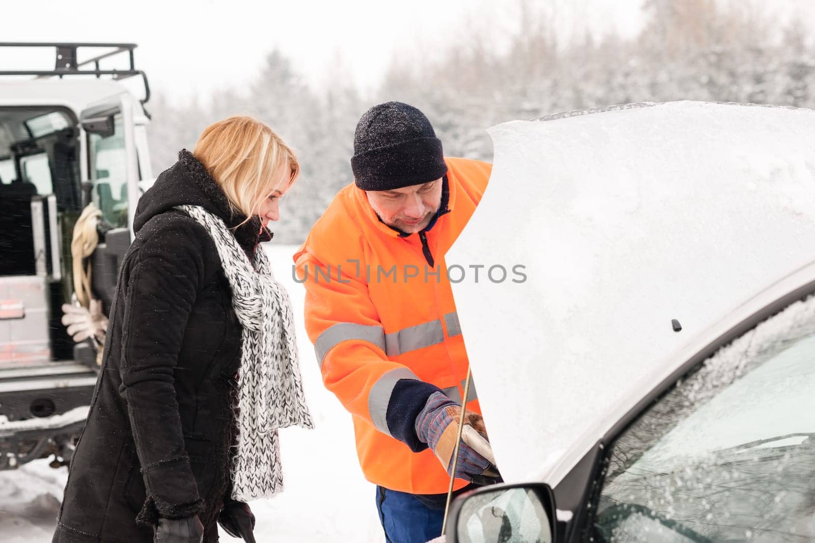 Woman mechanic looking under car hood snow assistance winter broken