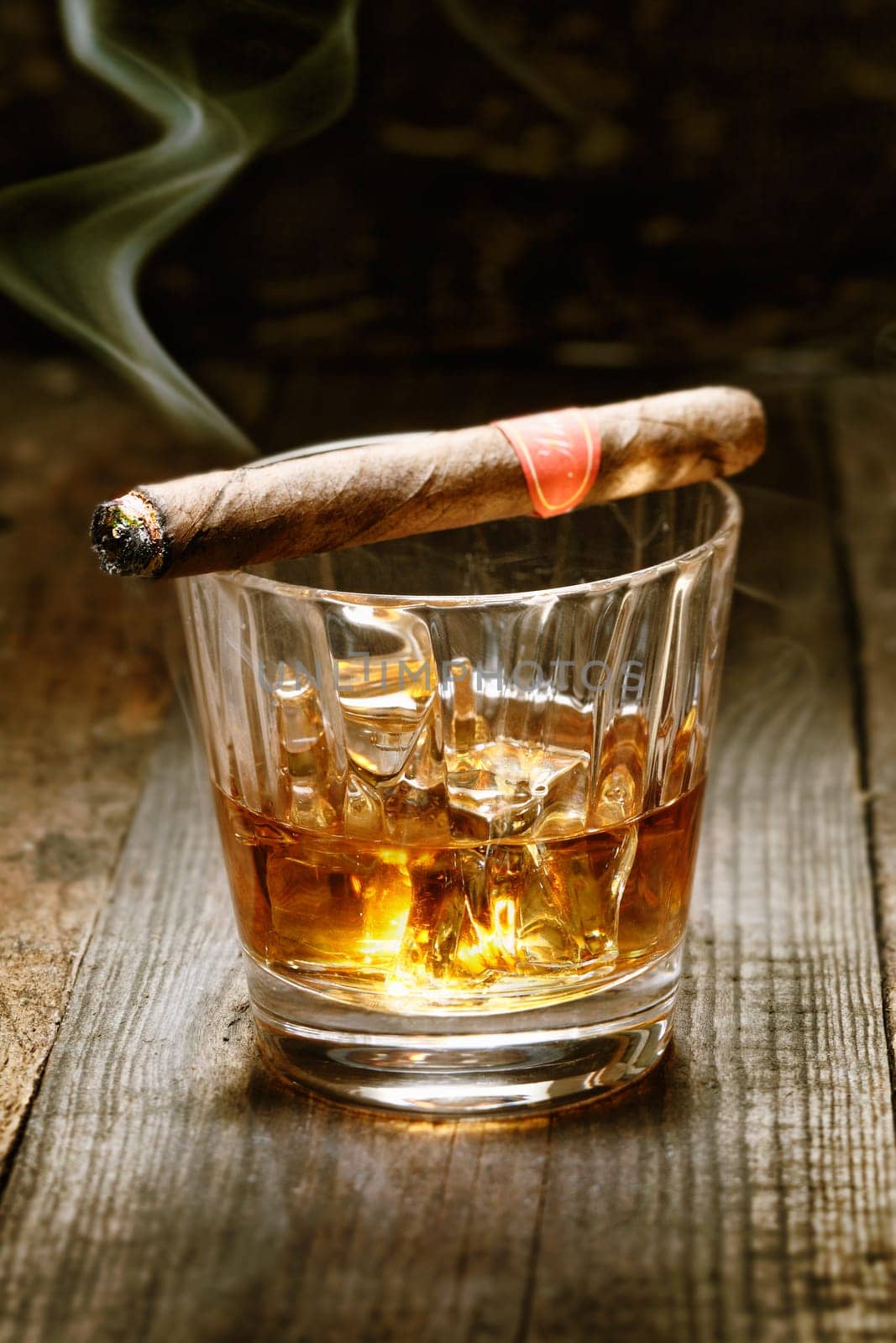Burning Cuban cigar resting on glass of whisky on ice on an old wooden surface symbolic of masculinity