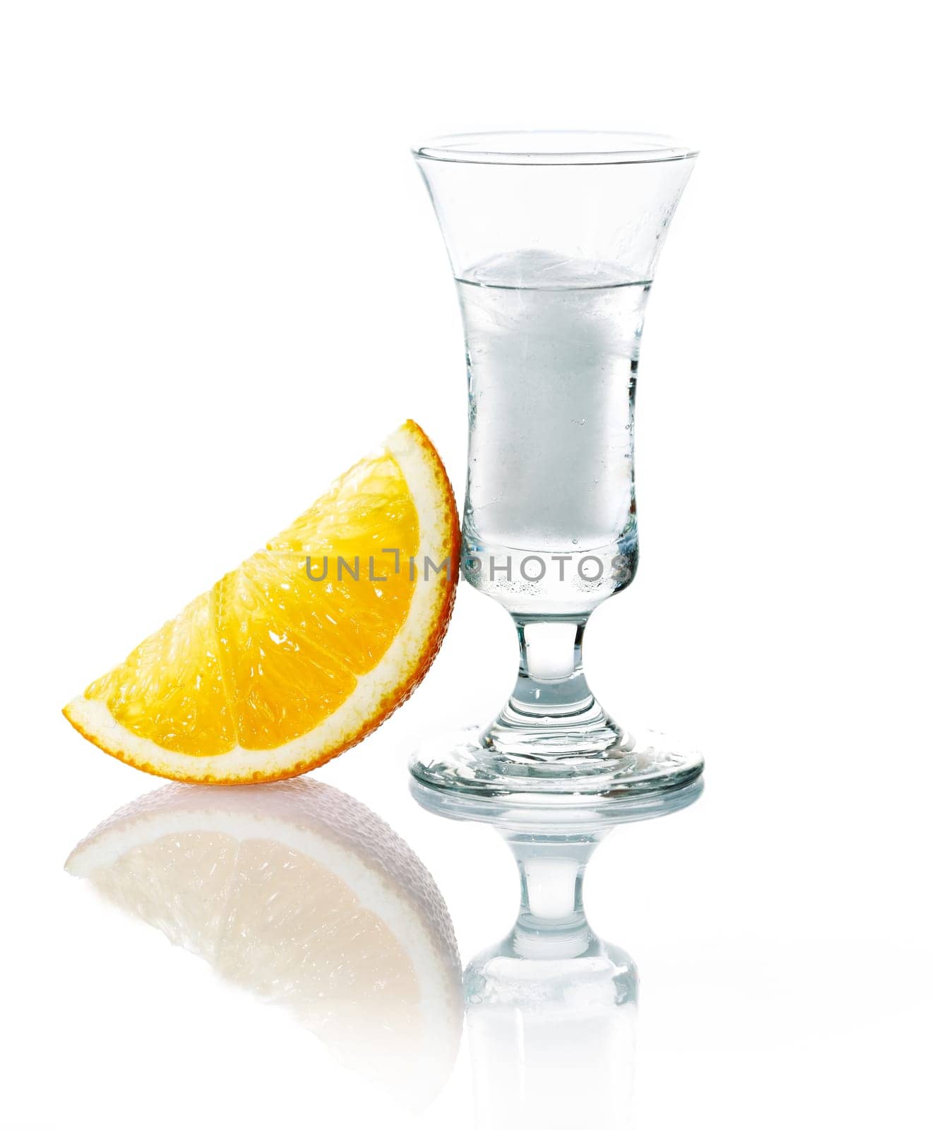 Shot glass with vodka or gin on the rocks served with a slice of fresh orange on a white studio background with reflection