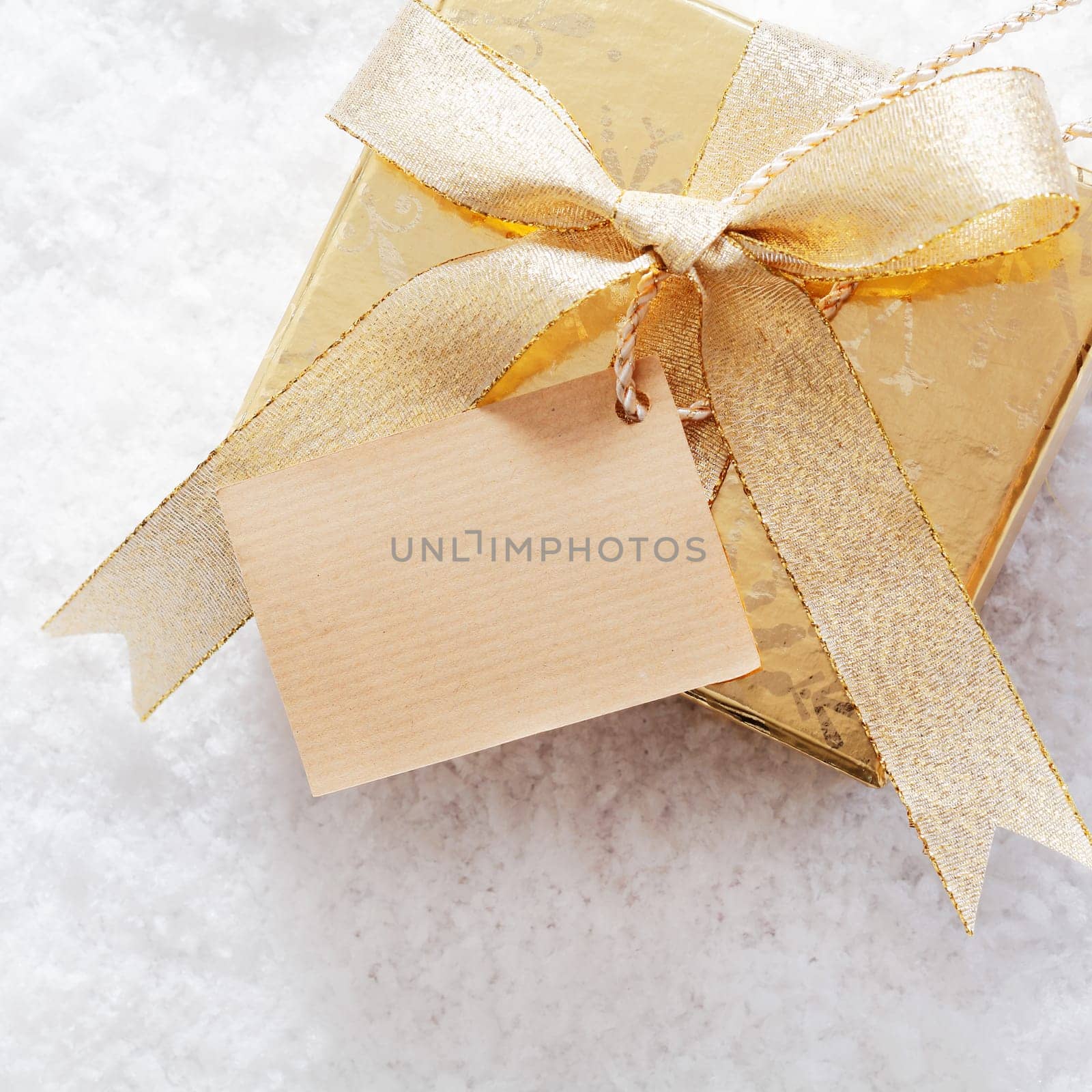 Gold Christmas giftbox with blank paper label and decorative ribbon and bow standing on a bed of fresh winter snow