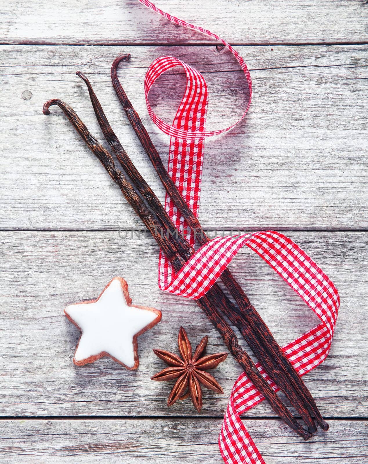 Country spices background with a colourful fresh red and white decorative ribbon twirled artistically through vanilla pods, star anise, and a Christmas star cookie on a weathered wood backgorund