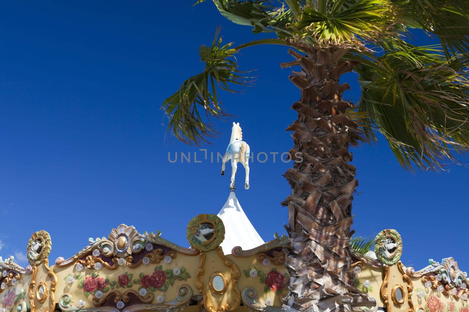 Beautiful colorful children's carousel with a peg on top stands behind a palm tree and blue sky in the marina of Gruissan in the south of France