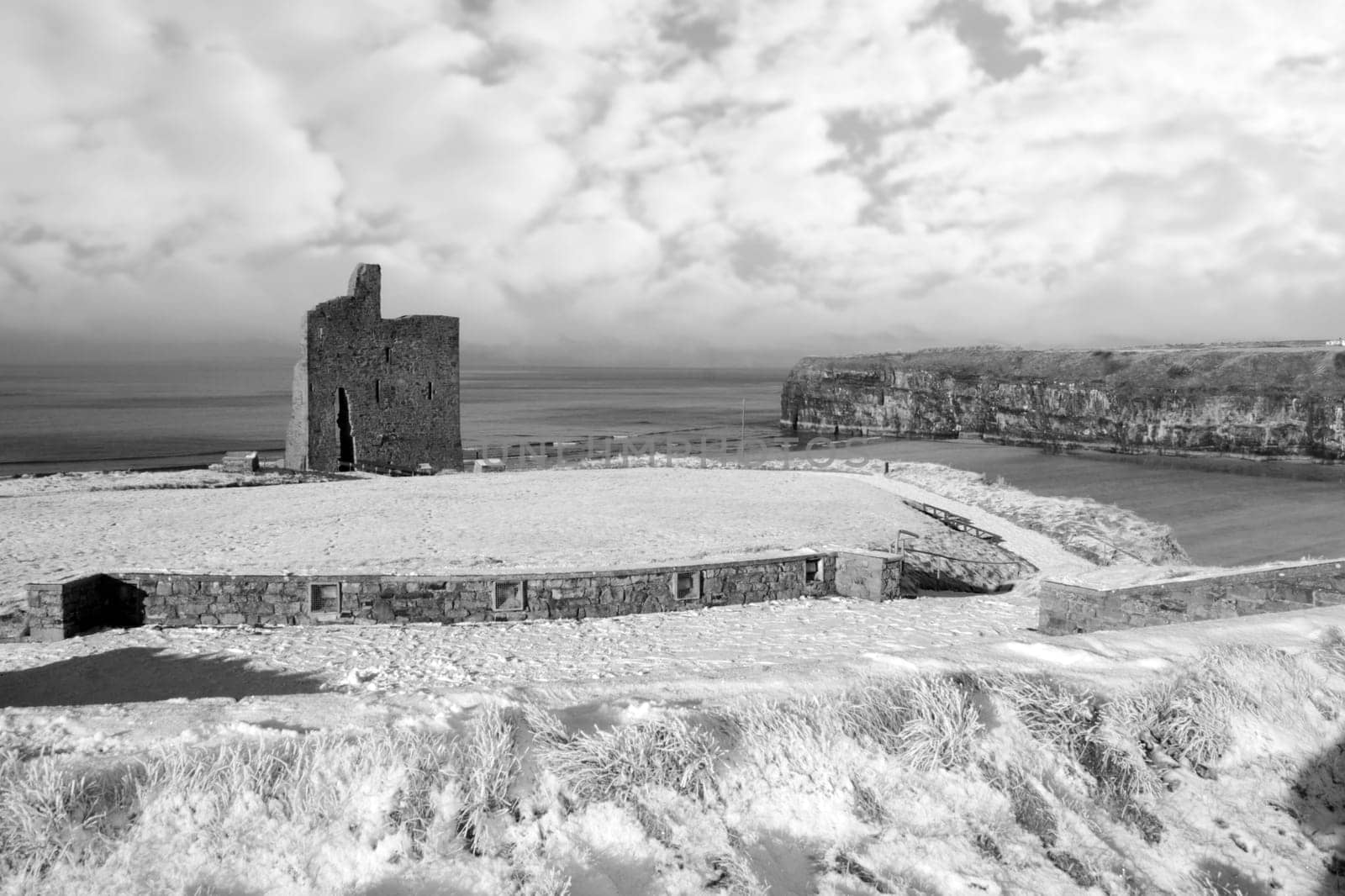 a seasonal snow covered view of atlantic ocean and ballybunion castle beach and cliffs on a frosty snow covered winters day in black and white