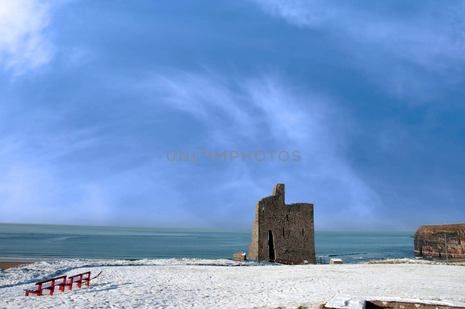 a seasonal snow covered view of atlantic ocean and ballybunion castle with red benches on a frosty snow covered winters day